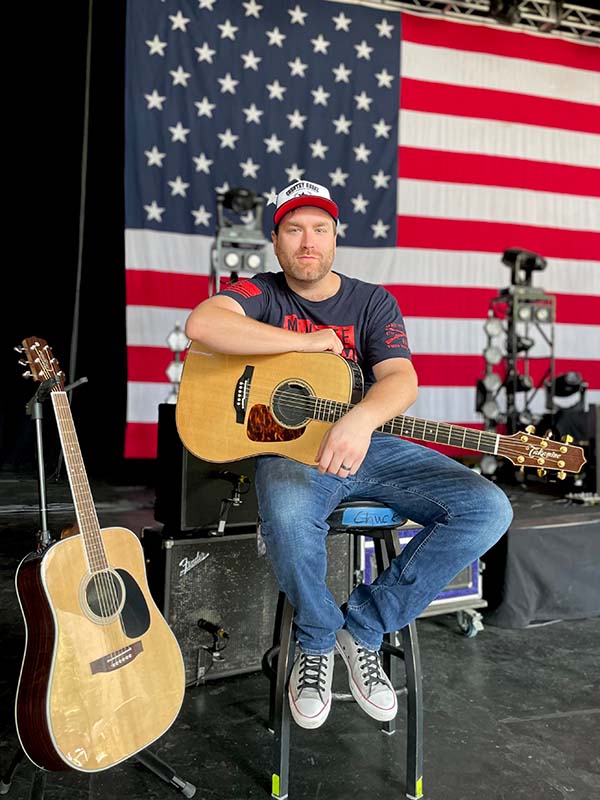 Chuck Ward holding his guitar with American flag behind him